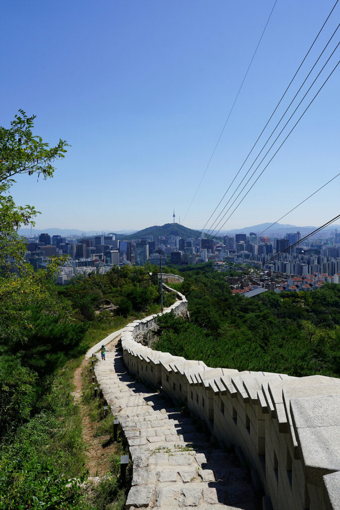 돌로 쌓은 성곽이 S자로 뻗어 있는 가운데 멀리 남산과 서울타워가 보인다. 하늘은 구름 한 점 없이 맑다.