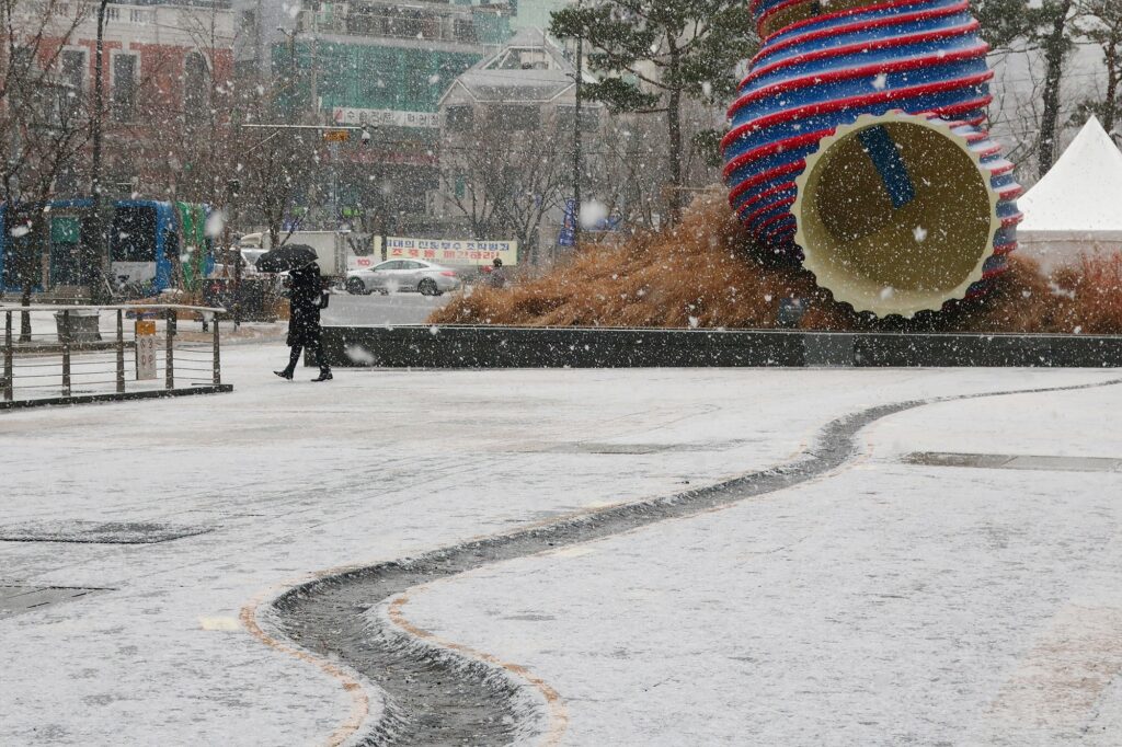 서울 청계광장에 눈이 내리고 있다. 검은 옷을 입은 행인 한 명이 검은 우산을 쓰고 소라기둥 앞을 지나가고 있다.