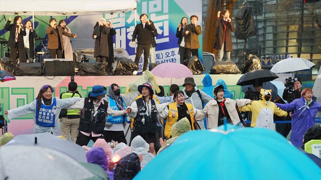 비가 내려 우산과 우비를 갖춘 시민들이 모여 있는 가운데, 무대 위와 무대 아래에 비상행동 측 공연자, 스태프, 연대자 등이 노래와 춤으로 승리의 기쁨을 나누고 있다.